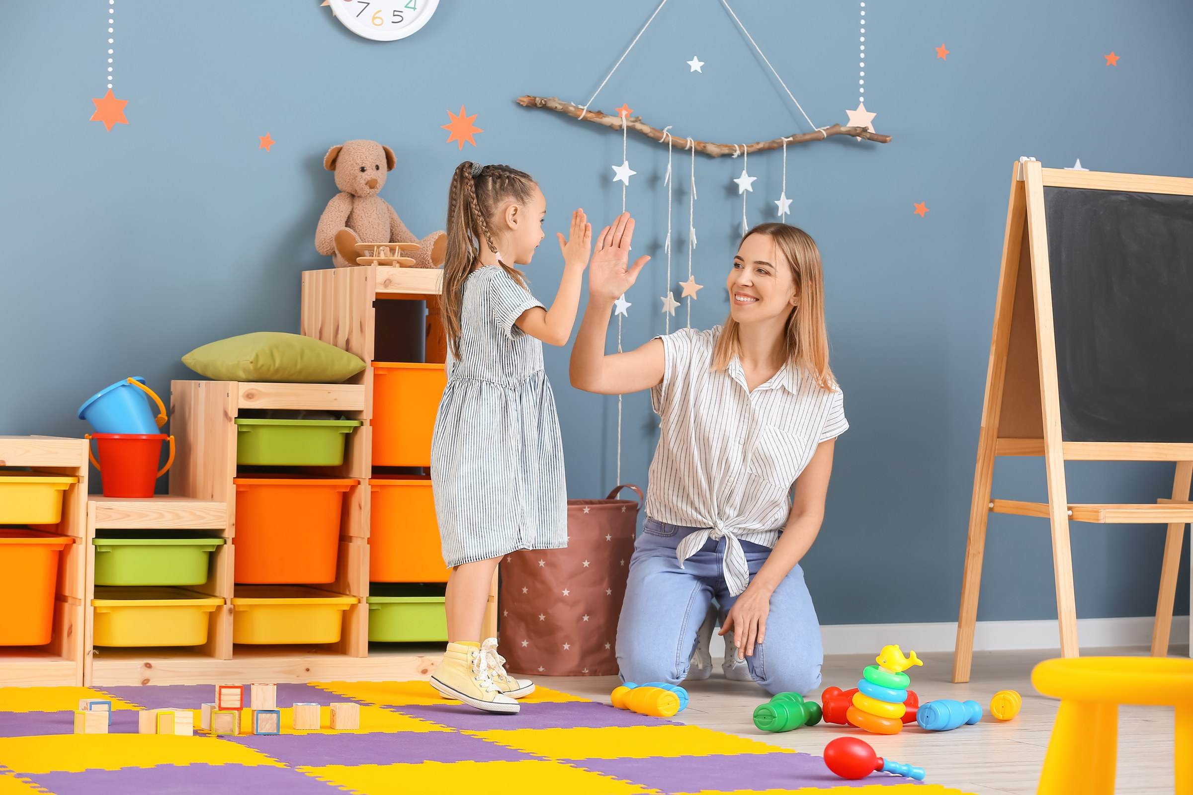 Eine Erzieherin und ein kleines Mädchen klatschen sich im bunten Spielzimmer ab.