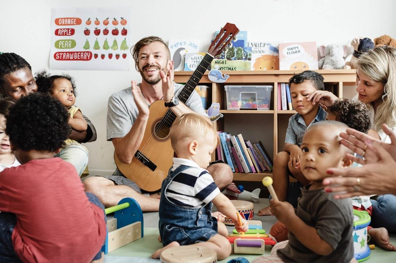 Kinder sitzen mit einem Erzieher mit Gitarre im Kreis und spielen auf Rhythmusinstrumenten.
