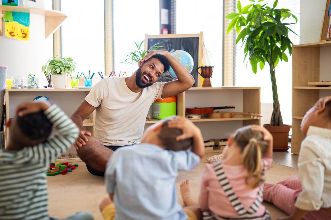 Ein Erzieher macht mit einer Kindergruppe auf dem Boden eine Bewegungsübung.